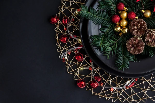 Christmas Decorations in the black plate on the dark background with some spruce twigs.Red, gold balls and cones inside and christmas light around . — стоковое фото