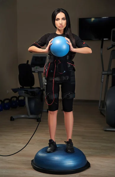 Brunette girl in EMS suit stands on bosu ball and holds blue bal