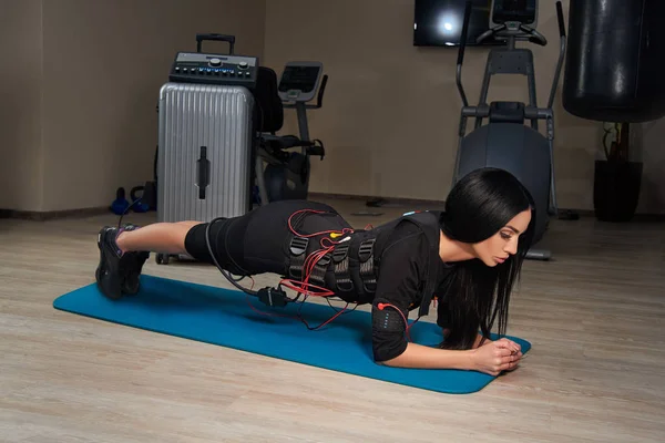 Beautiful brunette girl in EMS suit doing plank exercise on spor