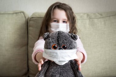 Coronavirus karantina konsepti, küçük kız COVID-19 salgını sırasında evde oyuncaklarla oynuyor. Korona virüsünü içeride korumak için maskeli bir çocuk. Coronavirüs nedeniyle evde kalan çocuk.