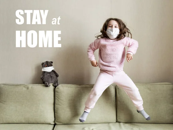 Evde karantinada kalmak, komik çocuk COVID-19 koronavirüs salgını sırasında koltuğa atlamak. Corona virüsünü korumak için maske takan küçük kız içeride oynuyor. Güvenli yaşam ve koronavirüs konsepti.