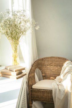 Pencerenin kenarındaki rahat okuma köşesinde rattan hasır sandalye, narin beyaz çigila çiçekleri, açık kitap ve pencere ışığı ve gölgeler. Modern rahat oturma odası alanı.