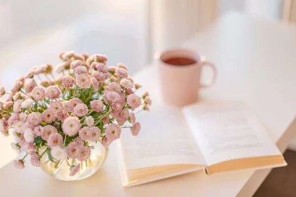 Cam vazoda küçük pembe çiçek buketi. Pembe çay ya da kahve fincanının bulanık odaklı arka planı ve pencerenin yanında açılan kitap..