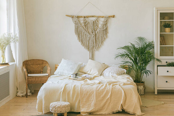 Modern romantic scandi boho style bedroom interior with decorative pillows, green plant and diy macrame wall panel. Light warm cozy comfortable home. jomo, weekend breakfast in bed concept.