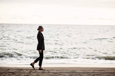 Siyah takım elbiseli güzel bir genç kız, eski moda, plajda yürüyor.