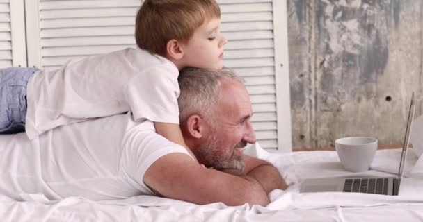 Pai e filho assistem desenhos animados em um laptop deitado na cama . — Vídeo de Stock