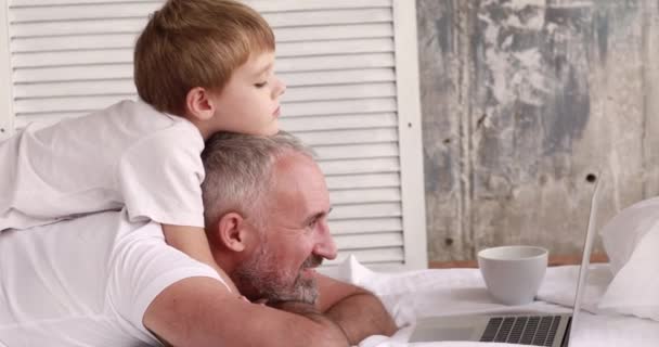 Apa és fia rajzfilmeket néznek egy laptopon, az ágyon fekve.. — Stock videók