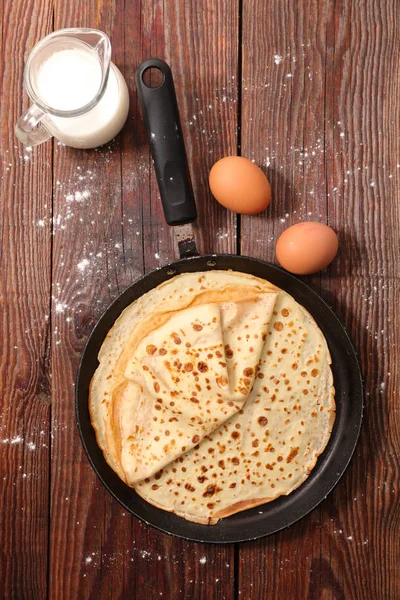 jug with milk and pan with crepes on wooden table