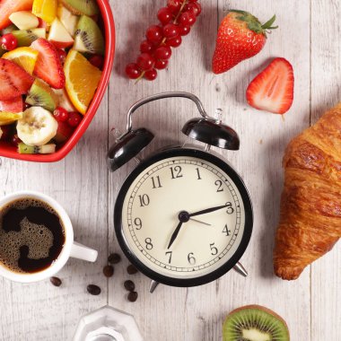 Çalar saat, meyve salatası, kruvasan ve kahve fincanı.