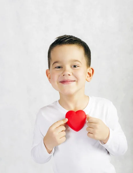 Ragazzo sorridente di cinque anni che tiene una statuetta rossa del cuore Immagine Stock