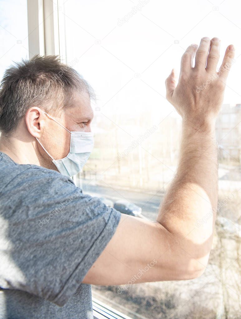 Sick sad man in medical protective face mask looking through window. Coronavirus pandemic covid-19 and quarantine concep