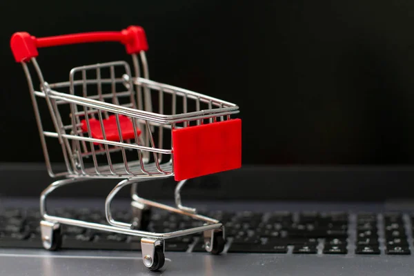 Pequeño carrito de compras en la computadora portátil de plata con fondo de pantalla negro. Compras online — Foto de Stock