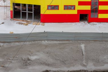 Yol kenarındaki bir çitin arkasındaki kış şehrinde inşaat halindeki cephe binaları.