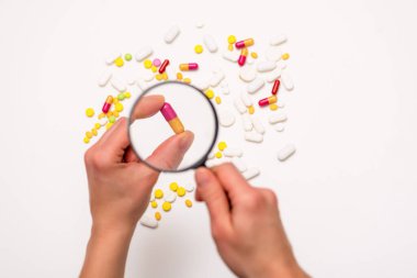 El büyüteç tutuyor. Beyaz arka plandaki farklı haplara büyüteçle bak. Seçici odaklanma. Sağlık, tıp ve bilim konsepti. Üst görünüm