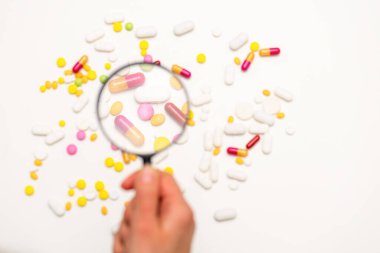 El büyüteç tutuyor. Beyaz arka plandaki farklı haplara büyüteçle bak. Seçici odaklanma. Sağlık, tıp ve bilim konsepti. Üst görünüm