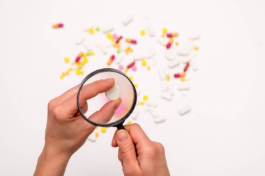 El büyüteç tutuyor. Beyaz arka plandaki farklı haplara büyüteçle bak. Seçici odaklanma. Sağlık, tıp ve bilim konsepti. Üst görünüm