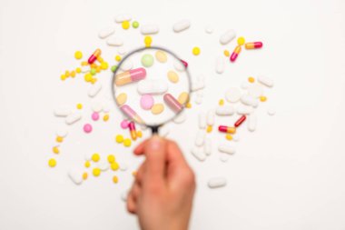 El büyüteç tutuyor. Beyaz arka plandaki farklı haplara büyüteçle bak. Seçici odaklanma. Sağlık, tıp ve bilim konsepti. Üst görünüm