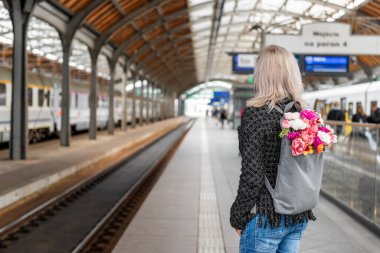 Bir buket çiçekli sırt çantalı genç kadın tren bekliyor. Fotoğraf arkadan..