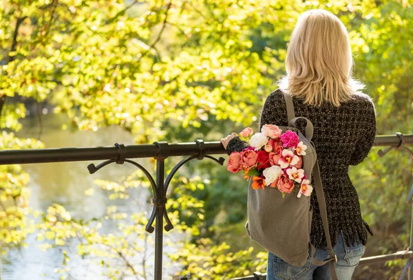 在春天的夏季公园里 年轻的金发美女背着一束花 背着灰色的背包 背对着照片 — 图库照片