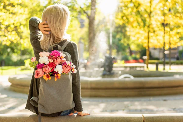 在春天的夏季公园里 年轻的金发美女背着一束花 背着灰色的背包 背对着照片 — 图库照片