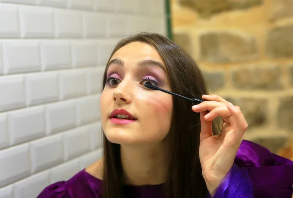Mujer morena aplicando maquillaje (pintarse las pestañas) delante de un espejo . — Foto de Stock