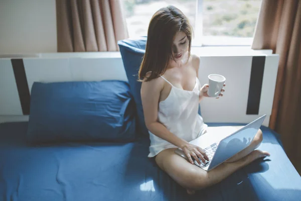 Mujer sosteniendo la taza y el uso de cuaderno en la cama . —  Fotos de Stock