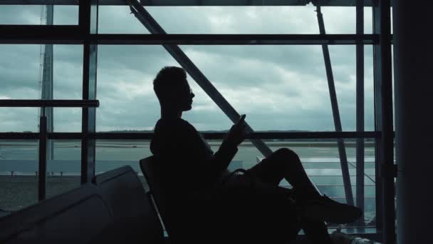 Silhouette eines Mannes, der am Flughafen Musik auf dem Smartphone hört — Stockvideo