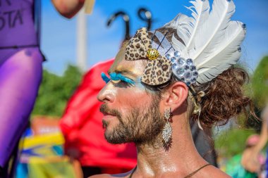 Transsexual with enormous blue eyelashes, earrings, headband with leopard bow and feathers, Bloco Orquestra Voadora, Carnaval 2017 clipart