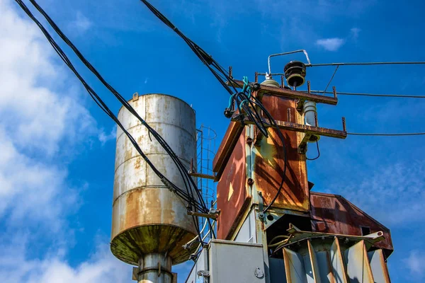 Industrial view of rusty transformer box, electrical wires and water tower with a ladder on the side
