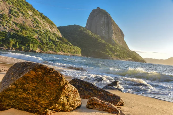 Große Steine am verlassenen Strand von Praia Vermelha und Sonnenaufgang mit der strahlenden Sonne, die den Zuckerhut erhellt — Stockfoto
