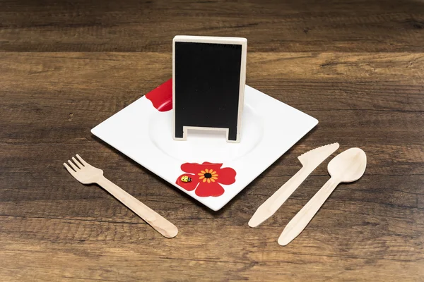 Small blackboard in white plate with spoon, fork and knife on wooden background — Stock Photo, Image