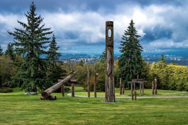 Burnaby Dağ Parkı 'ndaki totem direkleri kozalaklı ağaçların yanındaki yeşil çimlerin üzerinde fırtınalı bir gökyüzüne karşı. Vancouver Ufukta şehir merkezinde
