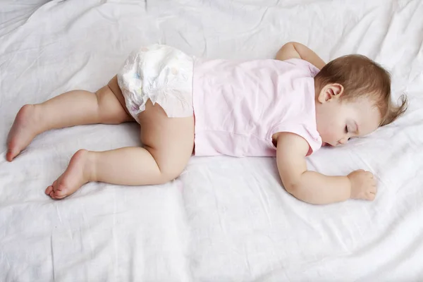 Egy pólós baba és egy pelenka egy fehér ágyon alszik a szemén. — Stock Fotó