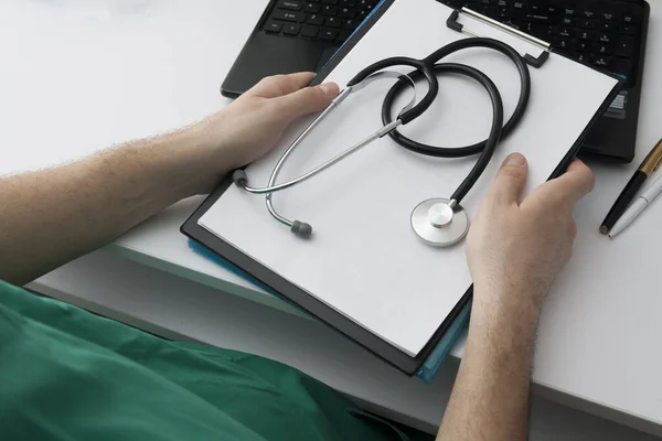 The doctor holds a tablet and a stethoscope in his hands over the table, behind the laptop. The workplace of a doctor. Medical concept — 스톡 사진