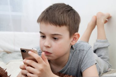 8-9 yaşlarında bir çocuk elinde telefonuyla yatakta yatıyor. Evde eğitim. Karantina koronavirüsü evde çocukları izole ediyor.