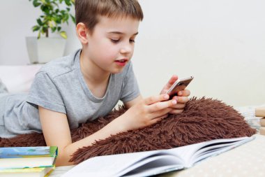 Bir çocuk elinde kitaplarla ve akıllı telefonuyla yatakta yatıyor. Evde eğitim. Karantina koronavirüsü evde çocukları izole ediyor.