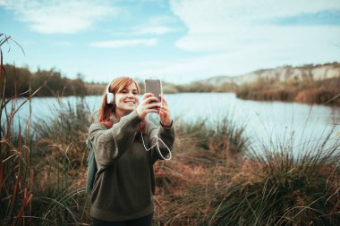 Genç bir kadın gölün kenarında akıllı telefonuyla selfie çekiyor.
