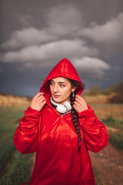 Junge Frau steht mit Regenmantel und Kopfhörer auf dem Feld — Stockfoto