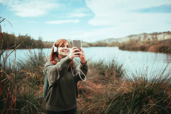 Young woman taking a selfie with her smartphone near a lake — Stock Photo, Image