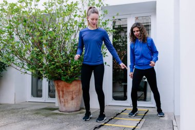 İki genç kadın terasta çeviklik merdiveni egzersizi yapıyor.