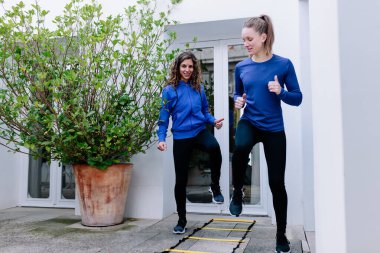 İki genç kadın terasta çeviklik merdiveni egzersizi yapıyor.