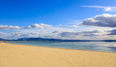 Sandy beach, sakin deniz, birkaç beyaz bulutlar arka plan ile mavi gökyüzü. Yaz hedef. Bulutlar ve dağlar deniz yansıması.