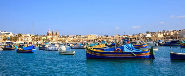 Malta, Marsaxlokk historiska hamn full av båtar. Blå himmel och byn bakgrund. Närbild av Visa, banner. — Stockfoto