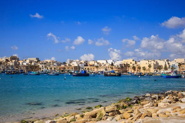 Marsaxlokk historic port with many boats in transparent sea and beach with stones, Malta. Blue sky and village background. — Stock Photo, Image