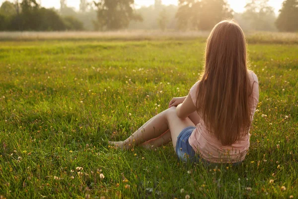Çimenlerin üzerinde oturan ve rahatlatıcı uzun siyah saçlı çekici bir genç kadın, arkadan görünüm — Stok fotoğraf