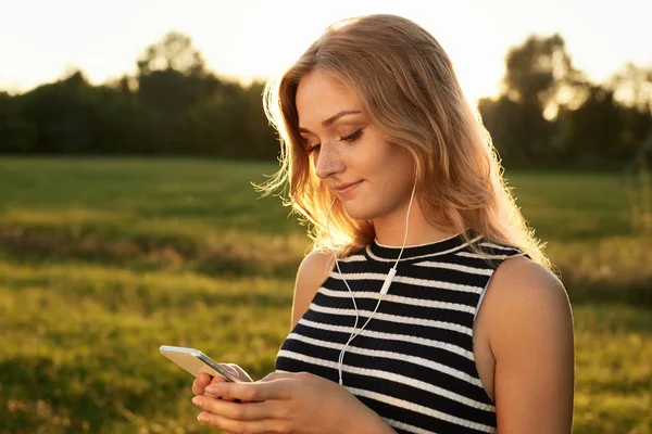 Portrait of a cute light-haired female who reads something on her smart phone and listens to the music with her earphones standing on the green background. Beautiful youg girl wearing striped T-shirt and holding her mobile phone — Stock Photo, Image