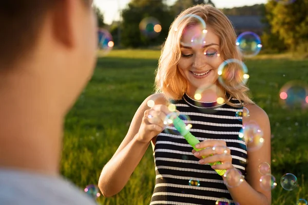 Krásná dívka se šťastným úsměvem foukání mýdlové bubliny na zelené louce se svým přítelem stojící zpět do fotoaparátu — Stock fotografie