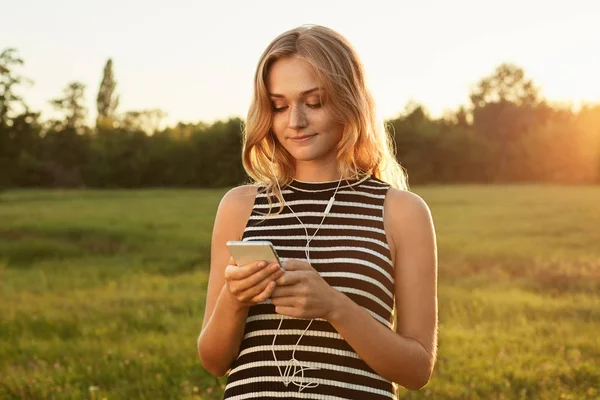 A happy girl with charming smile looking in her mobile phone and typing something. A walk on the green field. — Stock Photo, Image