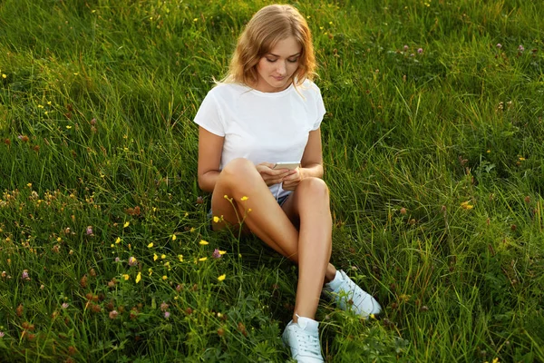 Menina atraente sentado de pernas cruzadas com seu telefone celular em uma grama vestindo uma camiseta branca e treinadores. Um olhar para um telemóvel — Fotografia de Stock