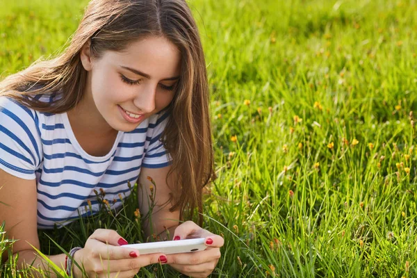Молодая девушка с длинными темными волосами и приятной улыбкой в полосатой футболке лежала на траве, держа в руках смартфон, печатая сообщения. Летнее время на лугу хорошая солнечная погода — стоковое фото